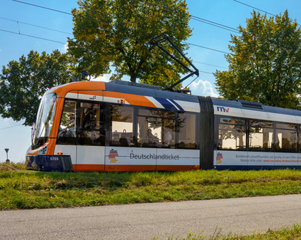 Eine Rhein-Neckar-Vario-Bahn (RNV-Bahn) fährt mit Werbung für das Deutschlandticket durch eine grüne Landschaft.