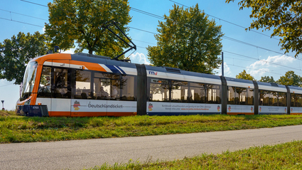 Eine Rhein-Neckar-Vario-Bahn (RNV-Bahn) fährt mit Werbung für das Deutschlandticket durch eine grüne Landschaft.