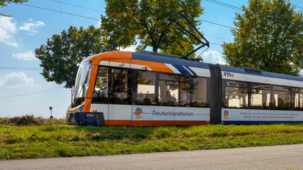 Eine Rhein-Neckar-Vario-Bahn (RNV-Bahn) fährt mit Werbung für das Deutschlandticket durch eine grüne Landschaft.