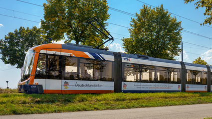 Eine Rhein-Neckar-Vario-Bahn (RNV-Bahn) fährt mit Werbung für das Deutschlandticket durch eine grüne Landschaft.