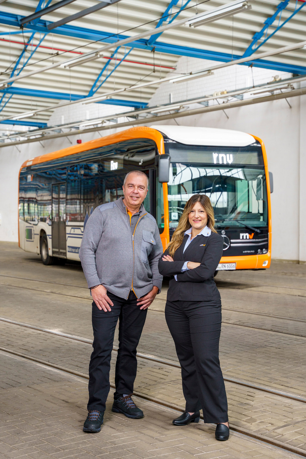 Eine Busfahrerin und ein Busfahrer der Rhein-Neckar-Verkehr GmbH (rnv) in Dienstkleidung vor einem Bus des Typs eCitaro in der Fahrzeughalle.